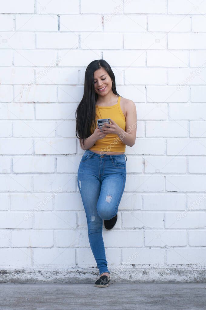 hermosa mujer latina escribiendo en su teléfono celular apoyada en una pared blanca
