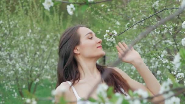 Весенний модный портрет красивой счастливой молодой женщины в белом платье с длинными каштановыми волосами бежать и наслаждаться запахом цветов в цветущем саду — стоковое видео
