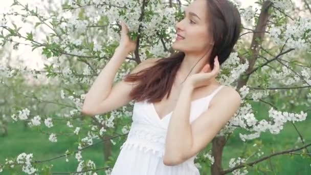 Retrato de moda de primavera de una hermosa mujer joven feliz en vestido blanco con pelo largo y marrón correr y disfrutar de flores de olor en el jardín floreciente — Vídeos de Stock