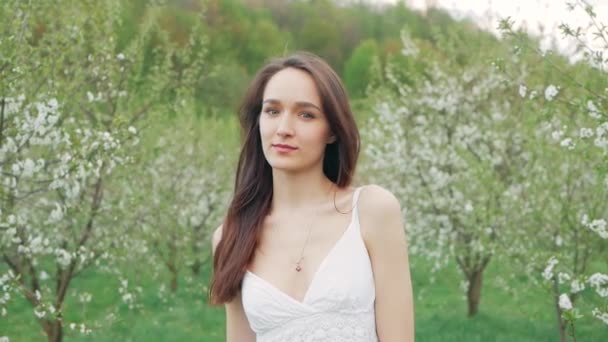 Retrato de moda de primavera de una hermosa mujer joven feliz en vestido blanco con pelo largo y marrón correr y disfrutar de flores de olor en el jardín floreciente — Vídeos de Stock
