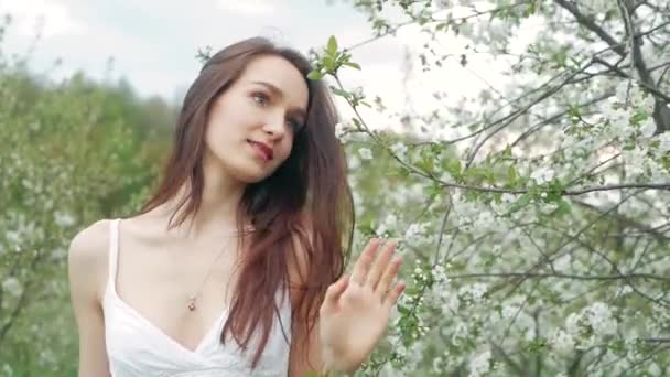 Retrato de moda de primavera de una hermosa mujer joven feliz en vestido blanco con pelo largo y marrón correr y disfrutar de flores de olor en el jardín floreciente — Vídeo de stock