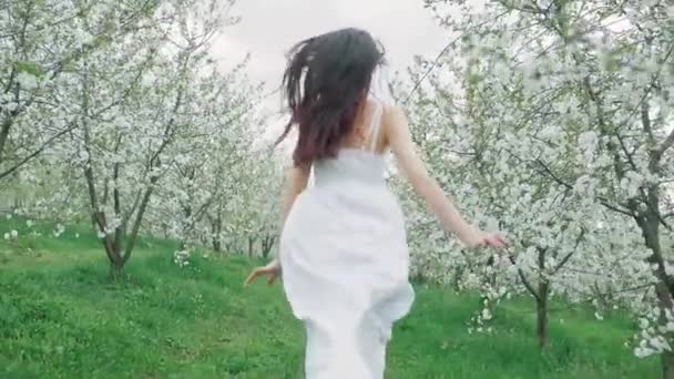 Άνοιξη μόδα πορτρέτο του μια όμορφη νεαρή ευτυχισμένη γυναίκα με λευκό φόρεμα με μακριά καστανά μαλλιά τρέχει και να απολαύσετε το ανθισμένο κήπο — Αρχείο Βίντεο