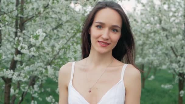 Retrato de moda de primavera de una hermosa mujer joven feliz en vestido blanco con pelo largo y marrón correr y disfrutar de flores de olor en el jardín floreciente — Vídeos de Stock