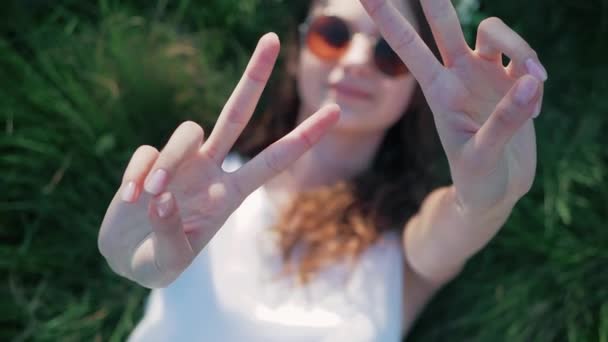 Slowmotion: Top view skott av en kaukasisk flicka i en vit blommig klänning liggande i gräset och visar ett tecken på fred — Stockvideo