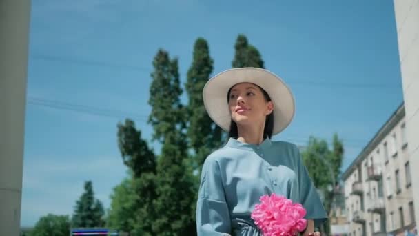 Счастливая стильная очаровательная молодая женщина смешанной расы Модный блоггер ходит по Европейской городской улице в синем летнем платье и белой шляпе и наслаждается отпускным приключением с розовыми цветами — стоковое видео
