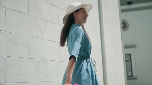 Happy Stylish Affascinante giovane donna di razza mista Fashion Blogger Camminando lungo European Urban Street in abito estivo blu e cappello bianco e godendo vacanza viaggio avventura whith fiori rosa — Video Stock