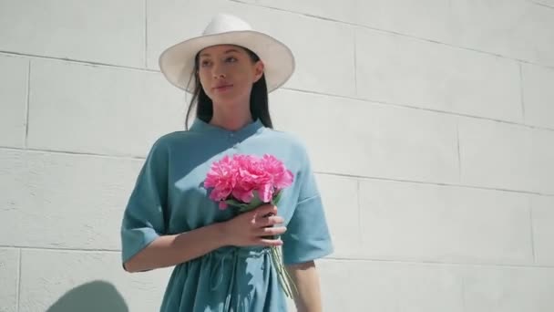幸せなスタイリッシュな魅力的な若い混合レース女性ファッションのブロガー歩行に沿ってヨーロッパ街路青い夏のドレスと白い帽子と楽しむ休日旅行休暇の冒険 whith ピンク花 — ストック動画