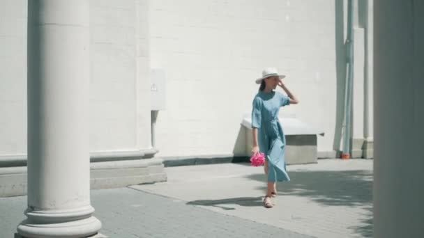 Happy elegant charmig ung blandad ras kvinna mode bloggare promenader längs Europeiska Urban Street i blå sommarklänning och vit hatt och njuter semester resa semester äventyr whith rosa blommor — Stockvideo