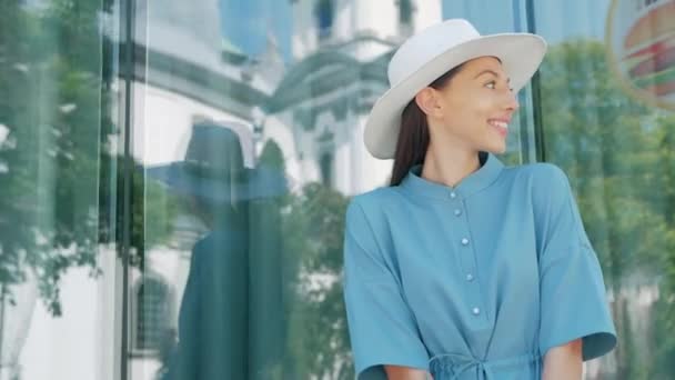 Retrato de Happy Stylish Encantadora joven mujer de raza mixta Blogger de moda en vestido azul de verano y sombrero blanco posando en la calle de verano, el sol brilla con flores rosadas — Vídeo de stock