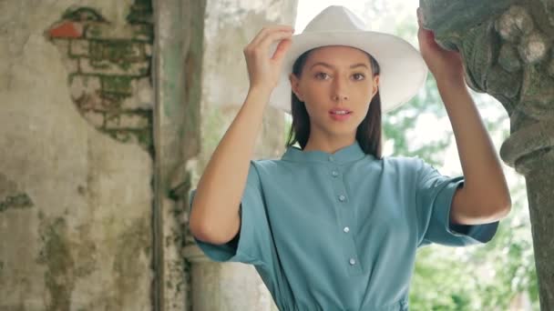 Retrato de feliz elegante encantadora joven mujer de raza mixta Blogger de moda en vestido azul de verano y sombrero blanco posando en la calle de verano, el sol está brillando — Vídeo de stock