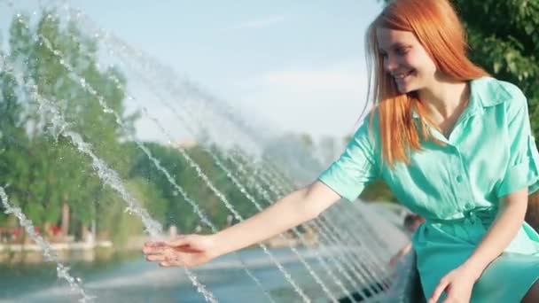 Menina com cabelo vermelho toca um fluxo de mão de água da fonte, câmera lenta — Vídeo de Stock