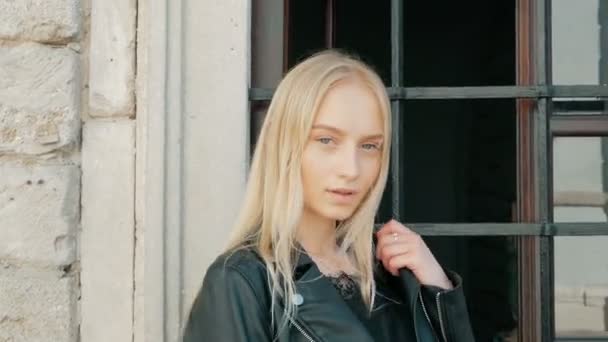 Portrait de mode d'une belle jeune femme aux yeux bleus et aux cheveux blonds dans une veste en cuir à l'extérieur — Video