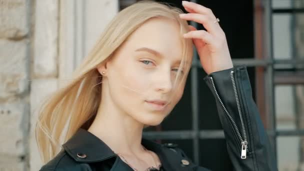 Portrait de mode d'une belle jeune femme aux yeux bleus et aux cheveux blonds dans une veste en cuir à l'extérieur — Video
