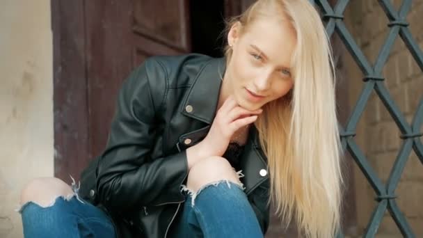Retrato de moda de una hermosa joven con ojos azules y cabello rubio en una chaqueta de cuero al aire libre — Vídeos de Stock