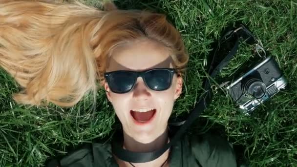 Vacker blond flicka ligger på gräset och dårar. tar bort och sätter på en hatt som liggande på gräset. begreppet fritid, semester, turism — Stockvideo