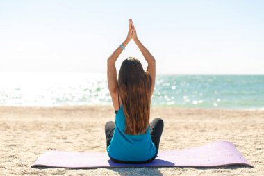 Siyah saçlı, beyaz bir kadın deniz sahilinde sabah meditasyonu yapıyor.