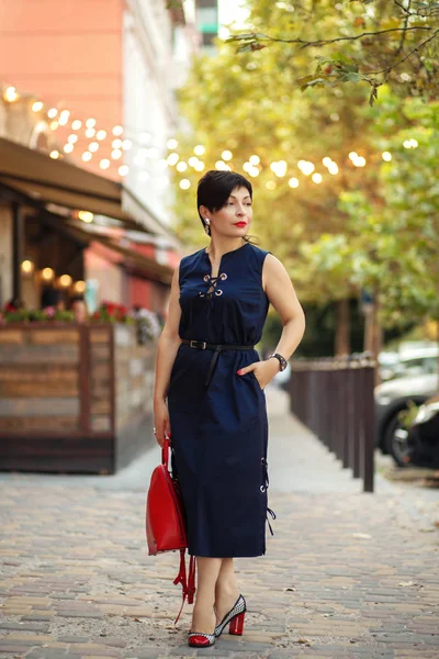 Outdoor portrait of short-cut brunette woman in stylish dress and red bag, trendy summer outfit posing in cafe, enjoying summer vacations