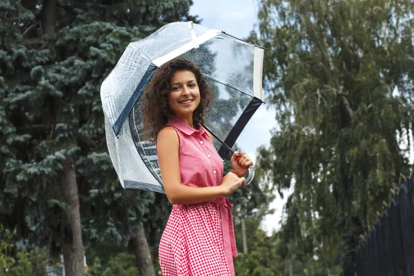 Young girl in a red dress with a transparent umbrella dancing in the rain standing in a puddle.