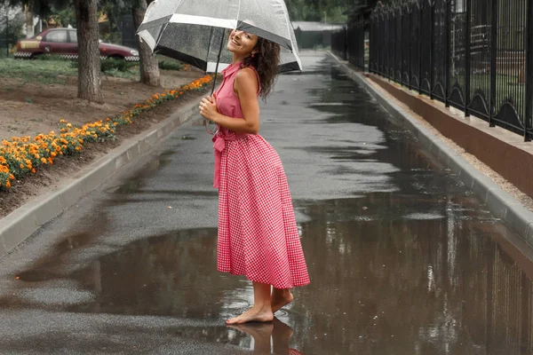 水たまりに立っている雨の中で踊る透明傘で赤いドレスを着た少女 — ストック写真
