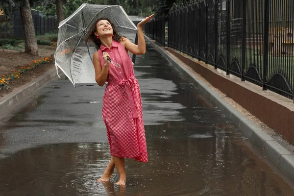 Young girl in a red dress with a transparent umbrella dancing in the rain standing in a puddle.