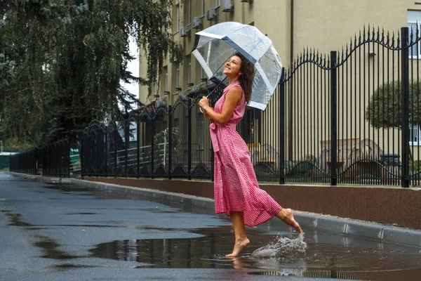 水たまりに立っている雨の中で踊る透明傘で赤いドレスを着た少女 — ストック写真