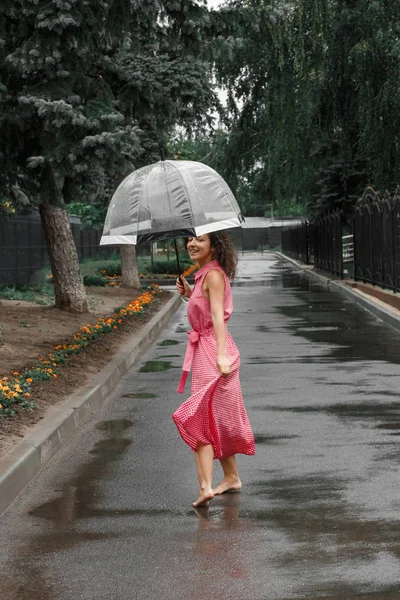 水たまりに立っている雨の中で踊る透明傘で赤いドレスを着た少女 — ストック写真