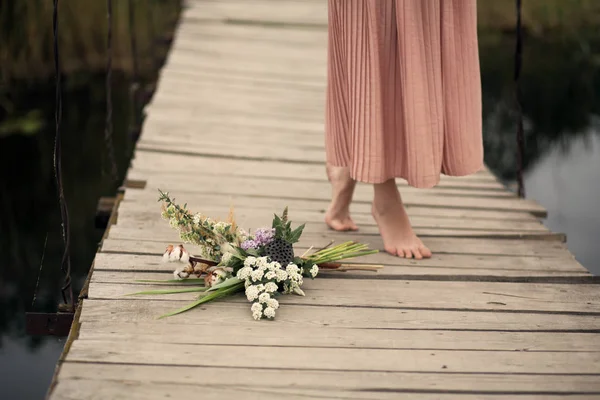 農村の木製の橋と花の花束に床を歩いて桃色ドレスの美しい柔らかい少女の足をクローズ アップ — ストック写真