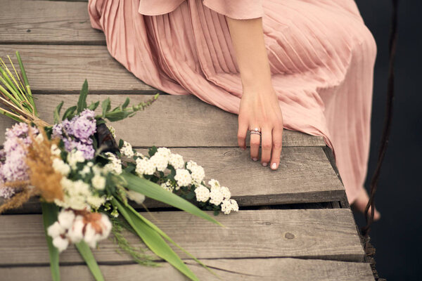 Beautiful tender girl in a peach-colored dress sits on a rural wooden bridge with a bouquet of flowers.
