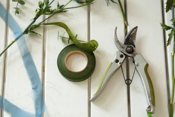 Pruner and other florist tools and cutted fresh flowers lying on white wooden table. Event fresh flowers decoration. Florist workflow. Wedding banquet design.