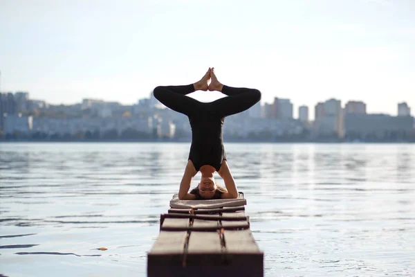 Young woman practicing yoga exercise at quiet wooden pier with city background. Sport and recreation in city rush.