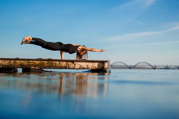 Young woman practicing yoga exercise at quiet wooden pier with city background. Sport and recreation in city rush.