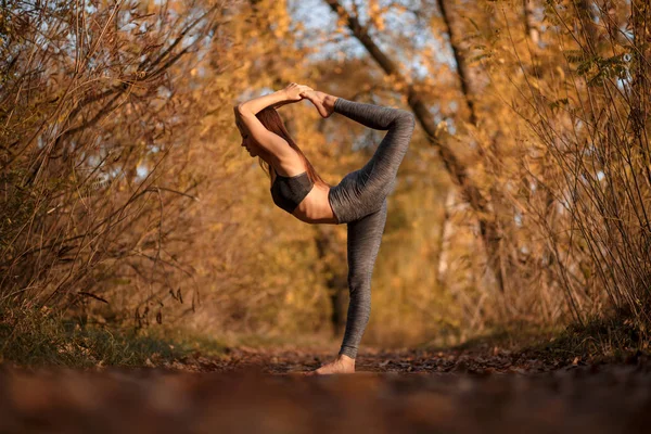 Young woman practicing yoga exercise in autumn park with yellow leaves. Sports and recreation lifestyle.