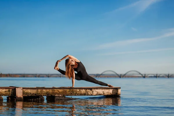 Young woman practicing yoga exercise at quiet wooden pier with city background. Sport and recreation in city rush.