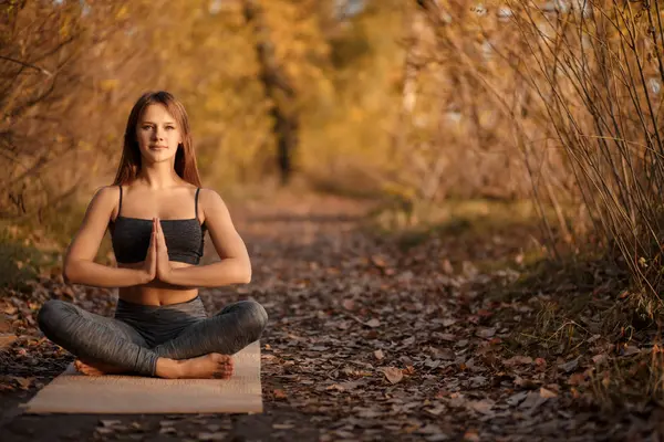 Young woman practicing yoga exercise in autumn park with yellow leaves. Sports and recreation lifestyle.