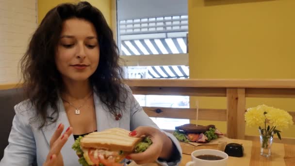 brünette Geschäftsfrau in blauer Jacke, isst ein frisches, üppiges Sandwich in einem gemütlichen Café mit gelben Wänden.