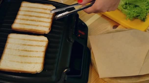 台所でサンドイッチを調理します トングが付いている手の上から見るサンドイッチ メーカーからトースト食パン パンを受け取り 紙ナプキンと木製のスタンドを産む — ストック動画