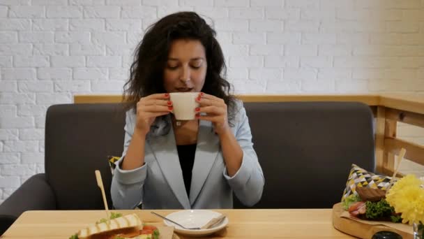 穿着蓝色夹克的肖像女孩 坐在灰色的沙发上吃早餐 喝了一口新鲜的咖啡 把杯子放在桌子上 把目光移开 — 图库视频影像