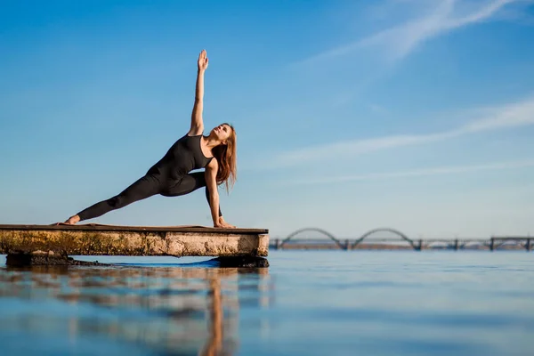 Young woman practicing yoga exercise at quiet wooden pier with city background. Sport and recreation in city rush.