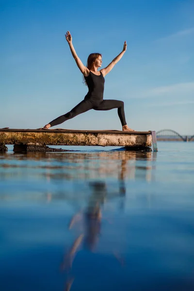 Young woman practicing yoga exercise at quiet wooden pier with city background. Sport and recreation in city rush.