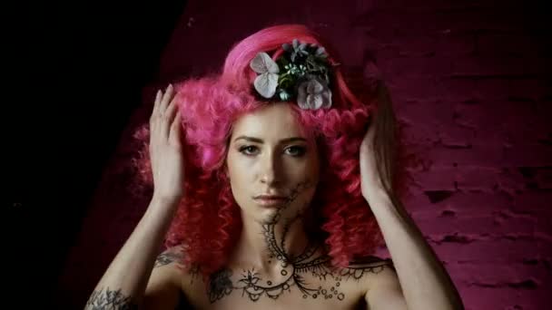 一个美丽的女孩与卷曲的粉红色头发时尚的发型的肖像 花编织成头发 在粉红色的墙壁的背景下 笑着和调情的微笑着对着镜头 — 图库视频影像