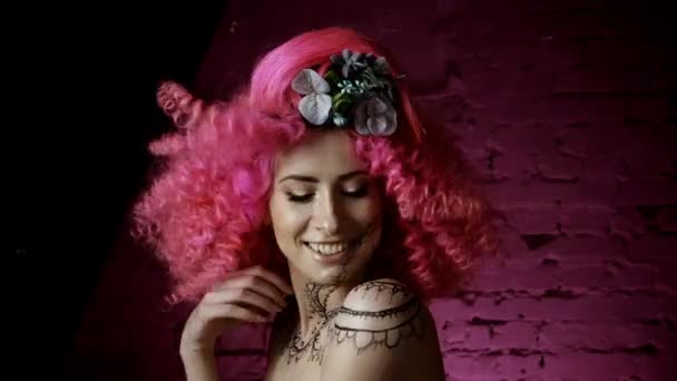 一个美丽的女孩与卷曲的粉红色头发时尚的发型的肖像 花编织成头发 在粉红色的墙壁的背景下 笑着和调情的微笑着对着镜头 — 图库视频影像