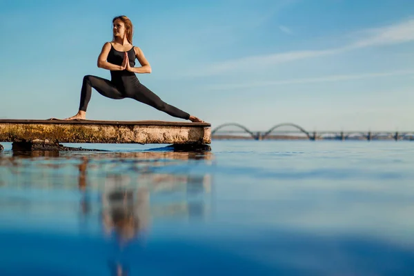 Young woman practicing yoga exercise at quiet wooden pier with city background. Sport and recreation in city rush.