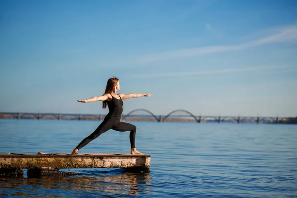 Young woman practicing yoga exercise at quiet wooden pier with city background. Sport and recreation in city rush.