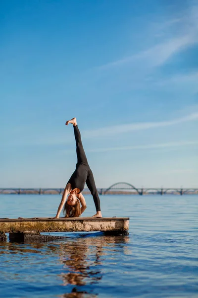 Young woman practicing yoga exercise at quiet wooden pier with city background. Sport and recreation in city rush.