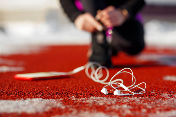 Спортсменка в черных кроссовках, приседающая на красной дорожке за побег. рядом находится телефон с проводными наушниками. Холодная снежная погода
.