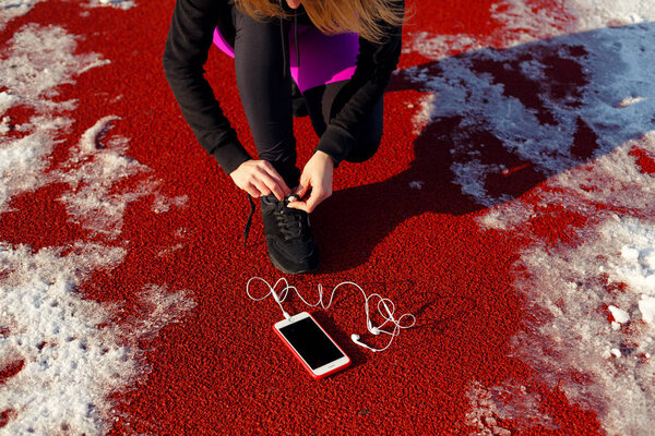 Спортсменка в черных кроссовках, приседающая на красной дорожке за побег. рядом находится телефон с проводными наушниками. Холодная снежная погода
.