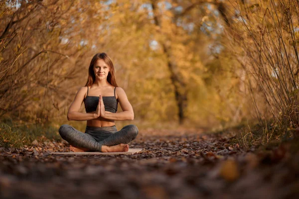 Young woman practicing yoga exercise at autumn park with yellow leaves. Sports and recreation lifestyle