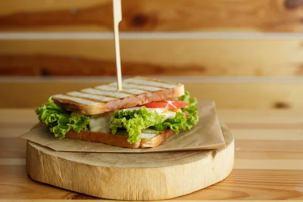 Juicy sandwich with bacon, fresh vegetables, green salad and dark lines after grill on wooden plate — Stock Photo, Image