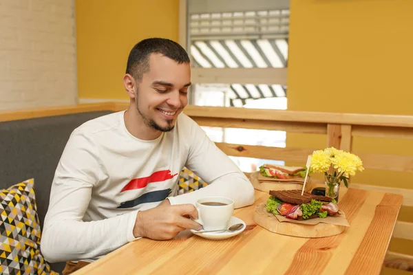 Jonge man glimlacht en nemen cup met koffie, Ontbijt in het café — Stockfoto