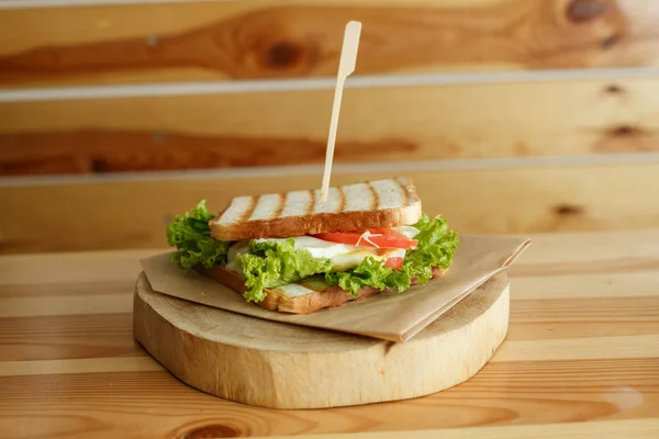 Сочный сэндвич с беконом, свежими овощами, зеленым салатом и темными линиями после гриля на деревянной тарелке — стоковое фото